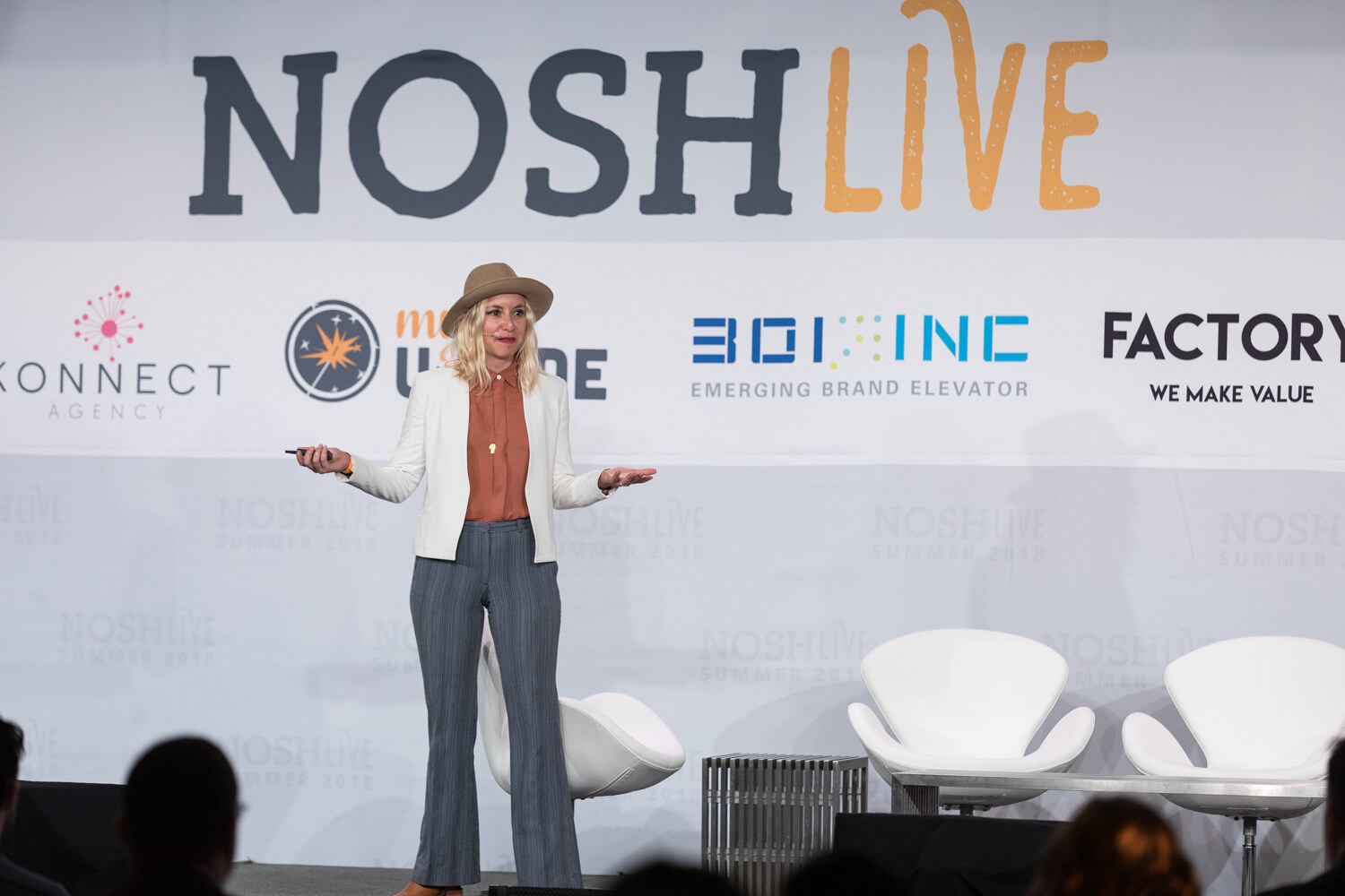 A presenter in a wide-brim hat and white blazer speaks on stage in front of the NOSH LIVE backdrop during the Build Your Badass Brand Voice session.