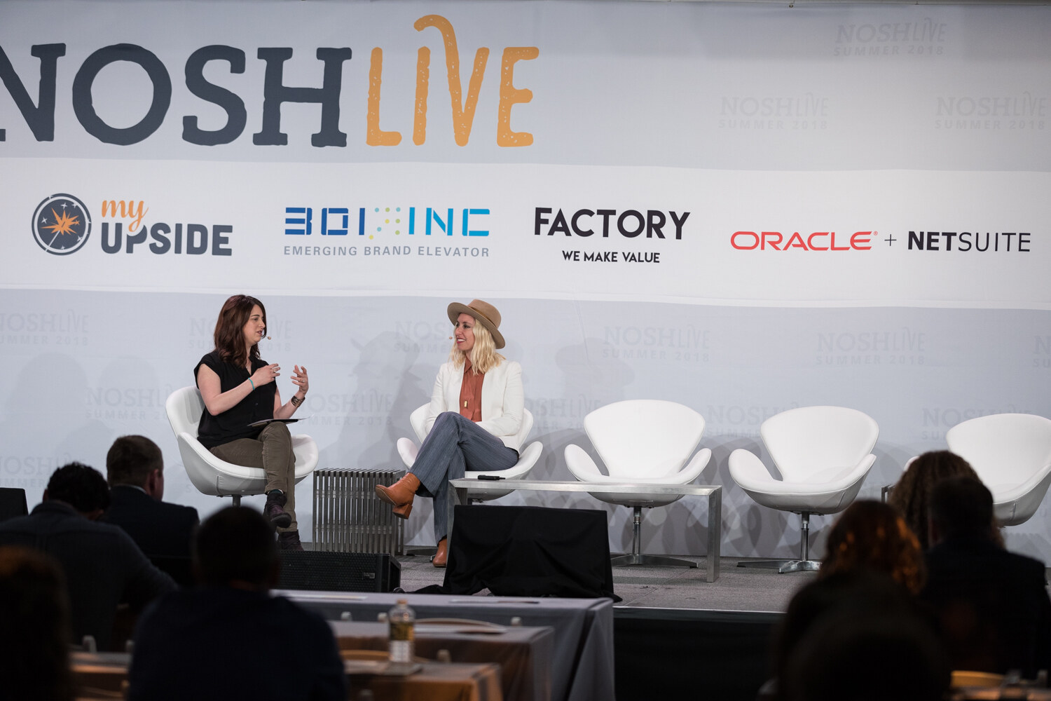 Two women sit in white chairs on stage during the Build Your Badass Brand Voice session with sponsor logos behind them.