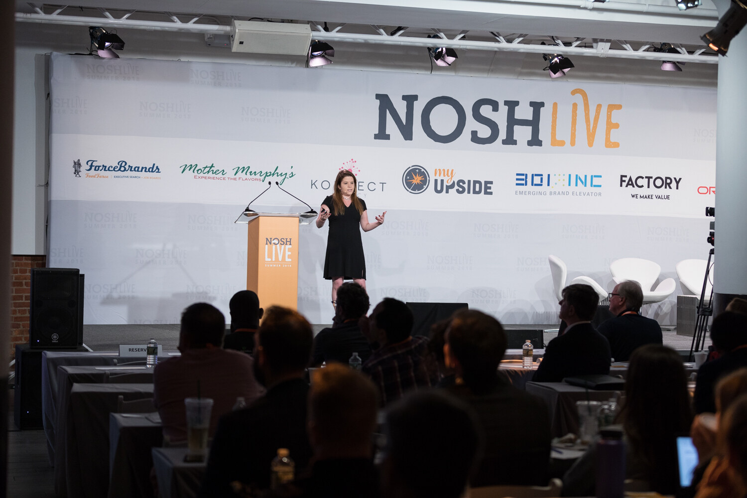 Wide view of a speaker on the Nosh Live stage with the audience in silhouette during The Investment Environment for Food Brands session.