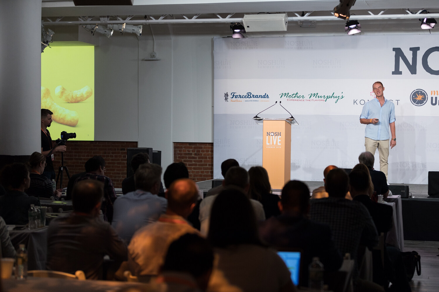 A presenter on stage beside the NOSH LIVE lectern with a yellow slide on a side screen during the Disrupting Big Categories session.