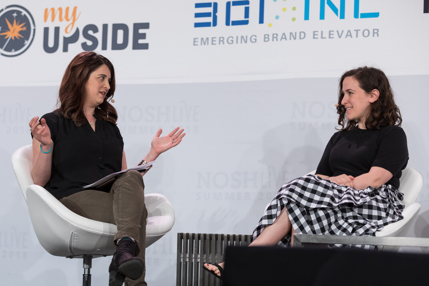 Two women in white chairs on stage, one gesturing mid-conversation, during the Feedback, Failing Fast, and Flying High in Direct to Consumer session.