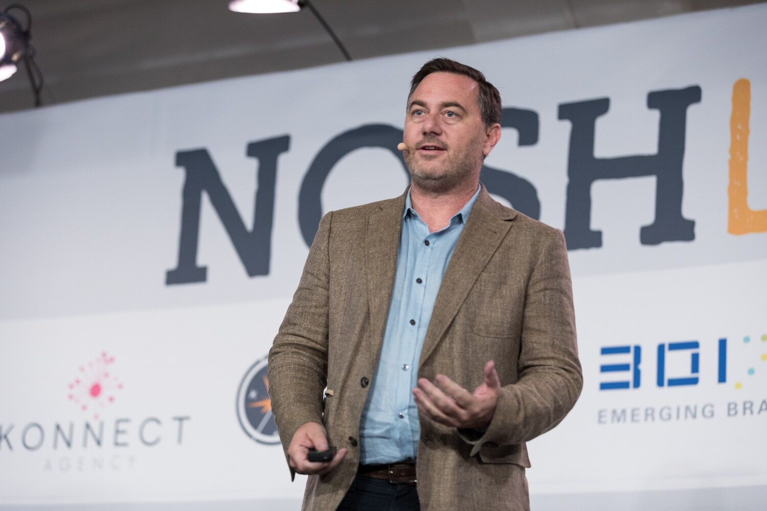 A presenter in a tweed blazer gestures in front of the NOSH LIVE backdrop during the Meal Kits, Brands, and Omnichannel session.