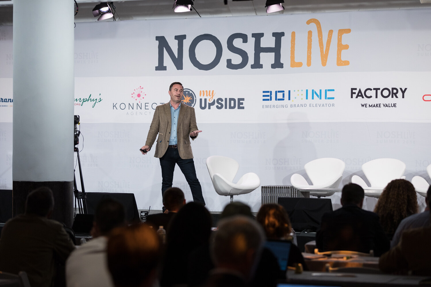 Wide view of a presenter on the Nosh Live stage with attendees and laptops in the foreground during the Meal Kits, Brands, and Omnichannel session.