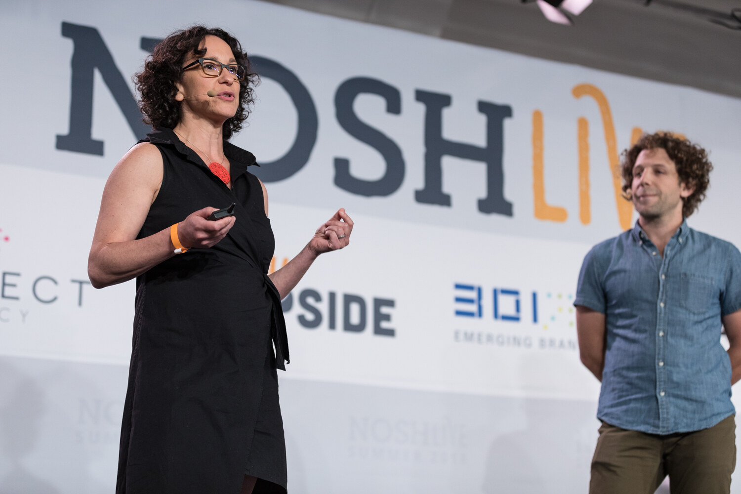 A presenter in a black dress speaks while her co-presenter stands beside her during The Move to Meaning: Product Becomes Brand session.