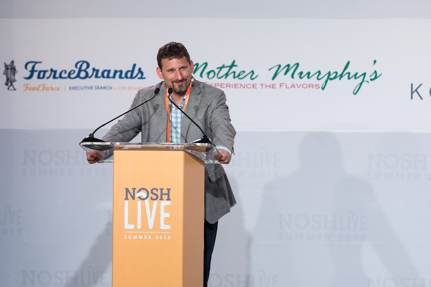 A speaker at the NOSH LIVE lectern opens the second day of the main conference with sponsor banners behind him.