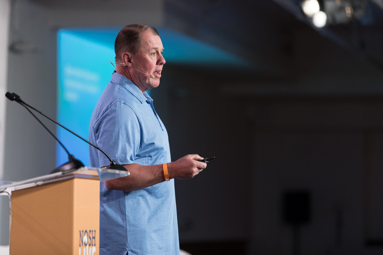A presenter in a light blue polo speaks beside the NOSH LIVE lectern during the Roles Meet Revenue session.