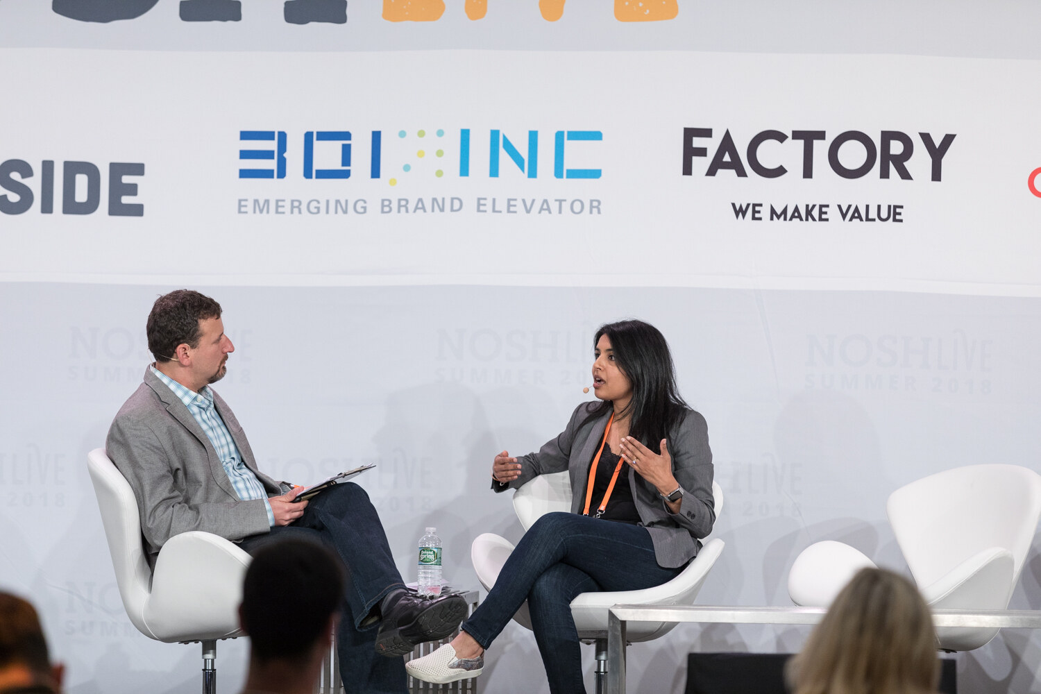 A moderator and a guest in a gray blazer sit in white chairs on stage during the From the Frontlines session.