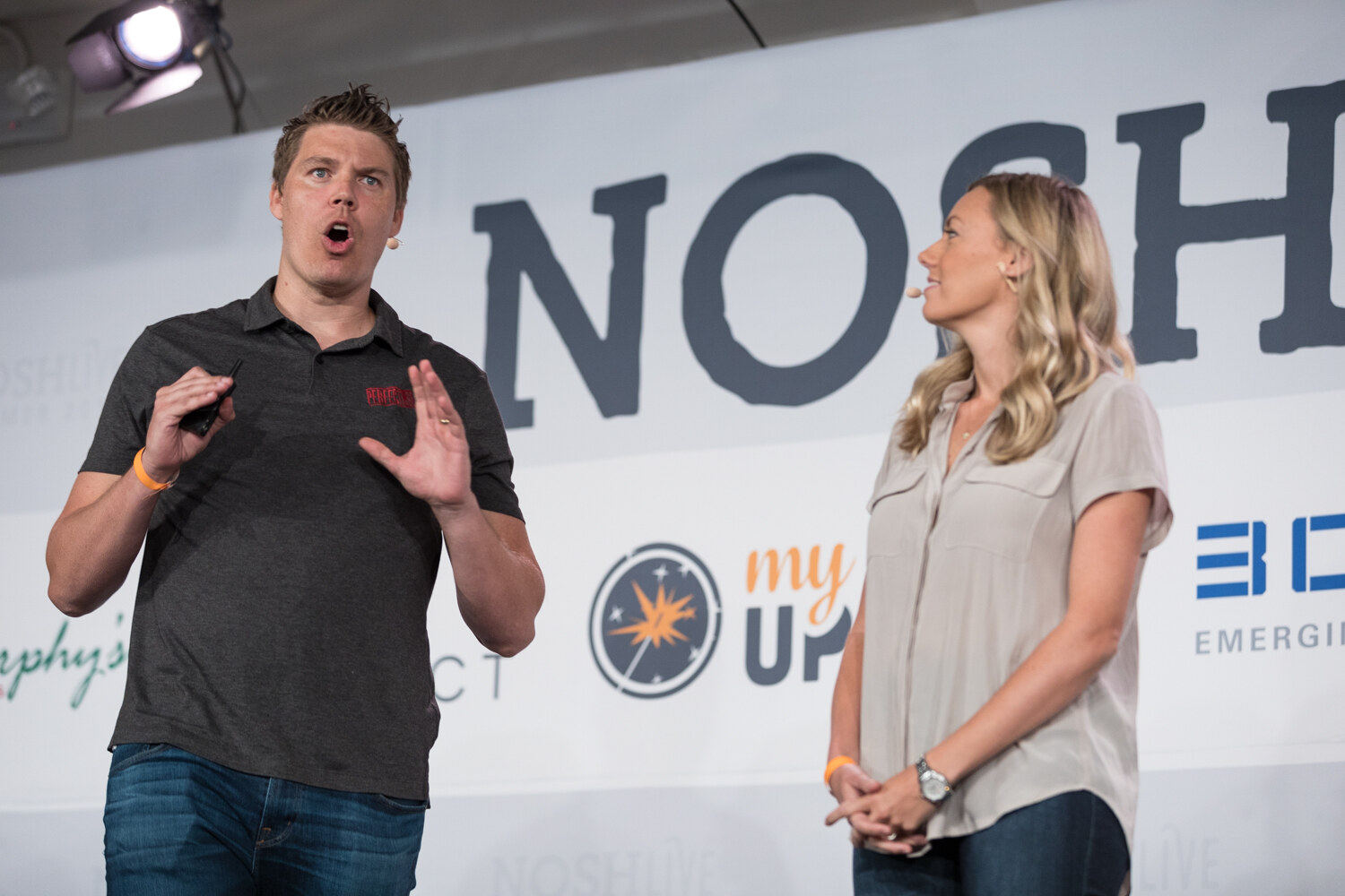 Two presenters, a man in a gray polo and a woman in a beige blouse, speak on stage during the New Category Development session.