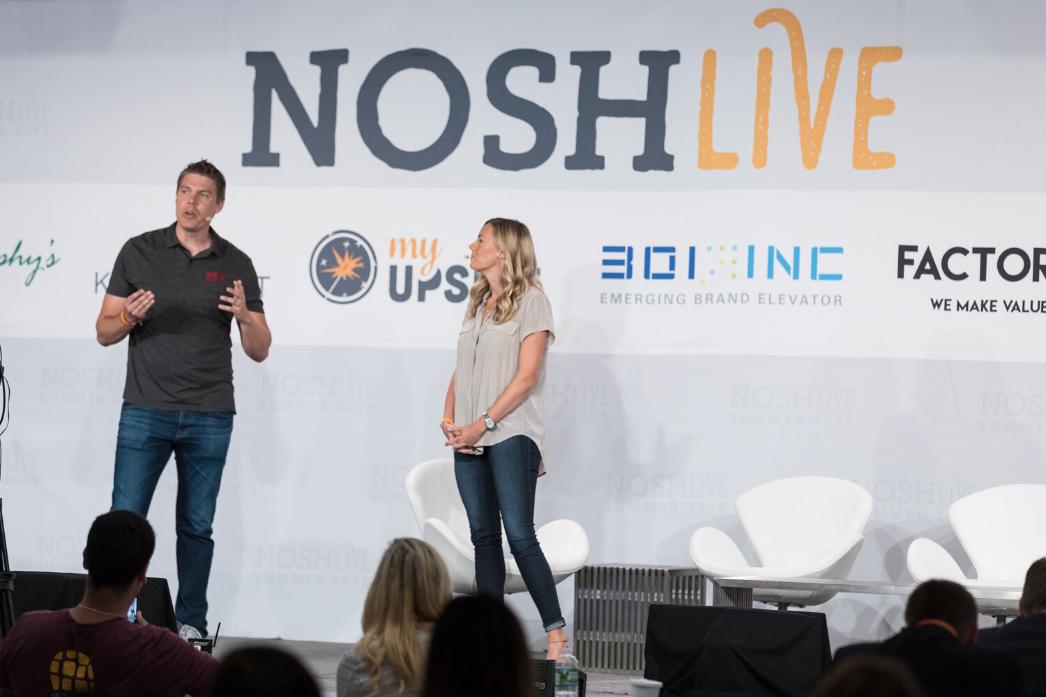 Wide view of two presenters on the Nosh Live stage with audience heads in the foreground during the New Category Development session.
