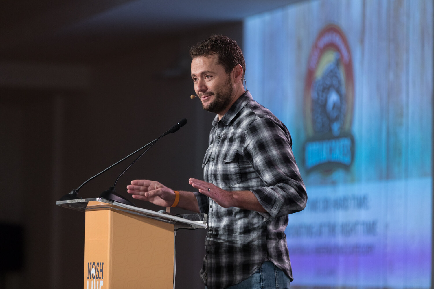 A presenter in a plaid shirt speaks at the lectern with a projected logo slide during the Hard Time session.