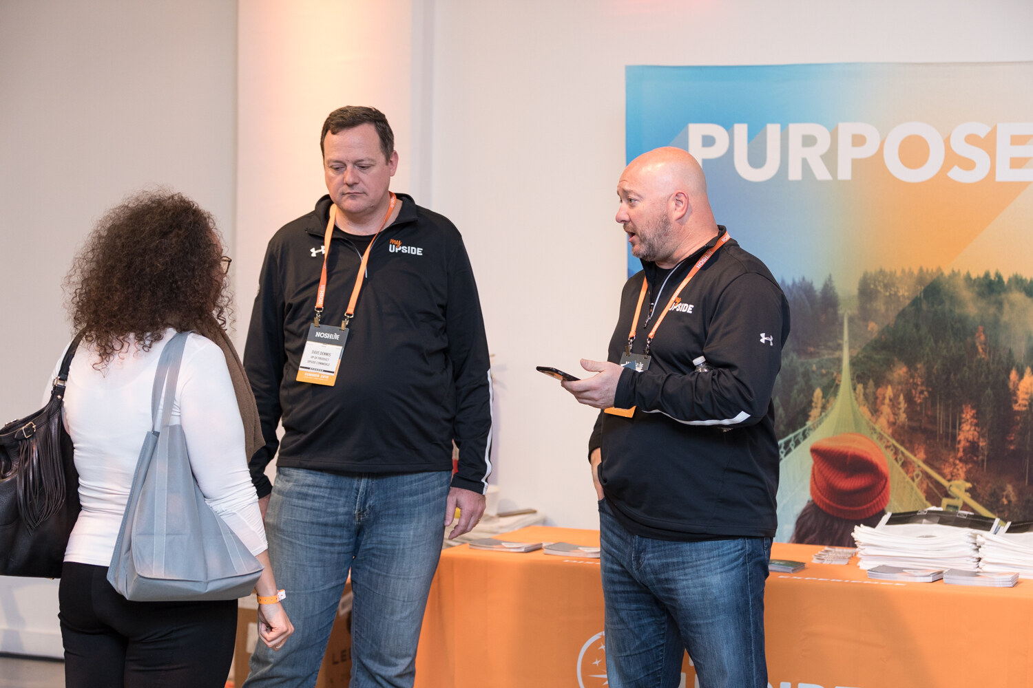 Two people in black pullovers stand at an orange sponsor table in front of a Purpose banner.