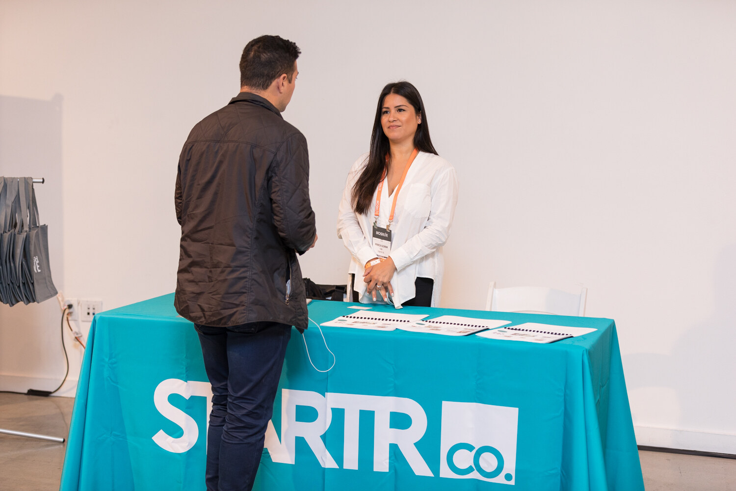 A woman in a white blazer talks with an attendee at a teal STARTR Co. table.