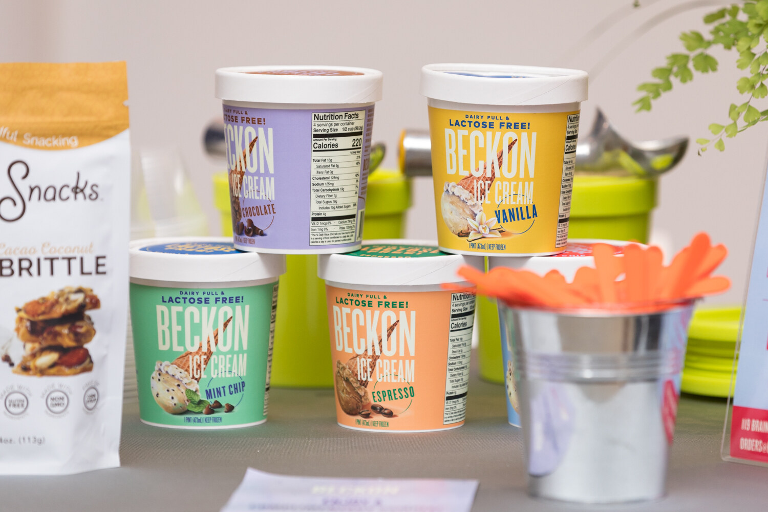 A sampling table of Beckon ice cream pints and a bag of Snacks brittle beside a pail of orange utensils.