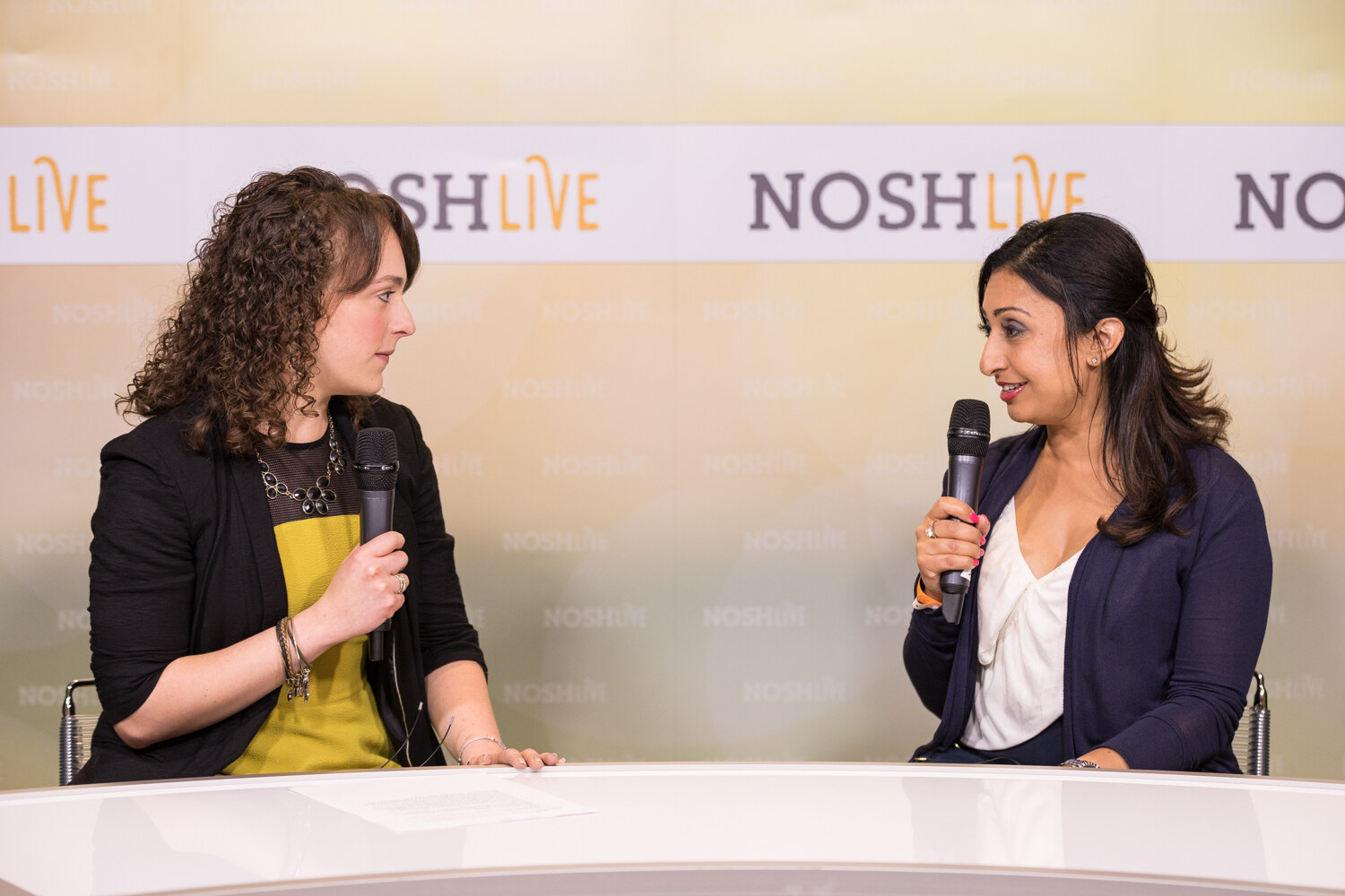 Two women with handheld microphones talk at the white Nosh Live livestream desk in front of a NOSH LIVE backdrop.