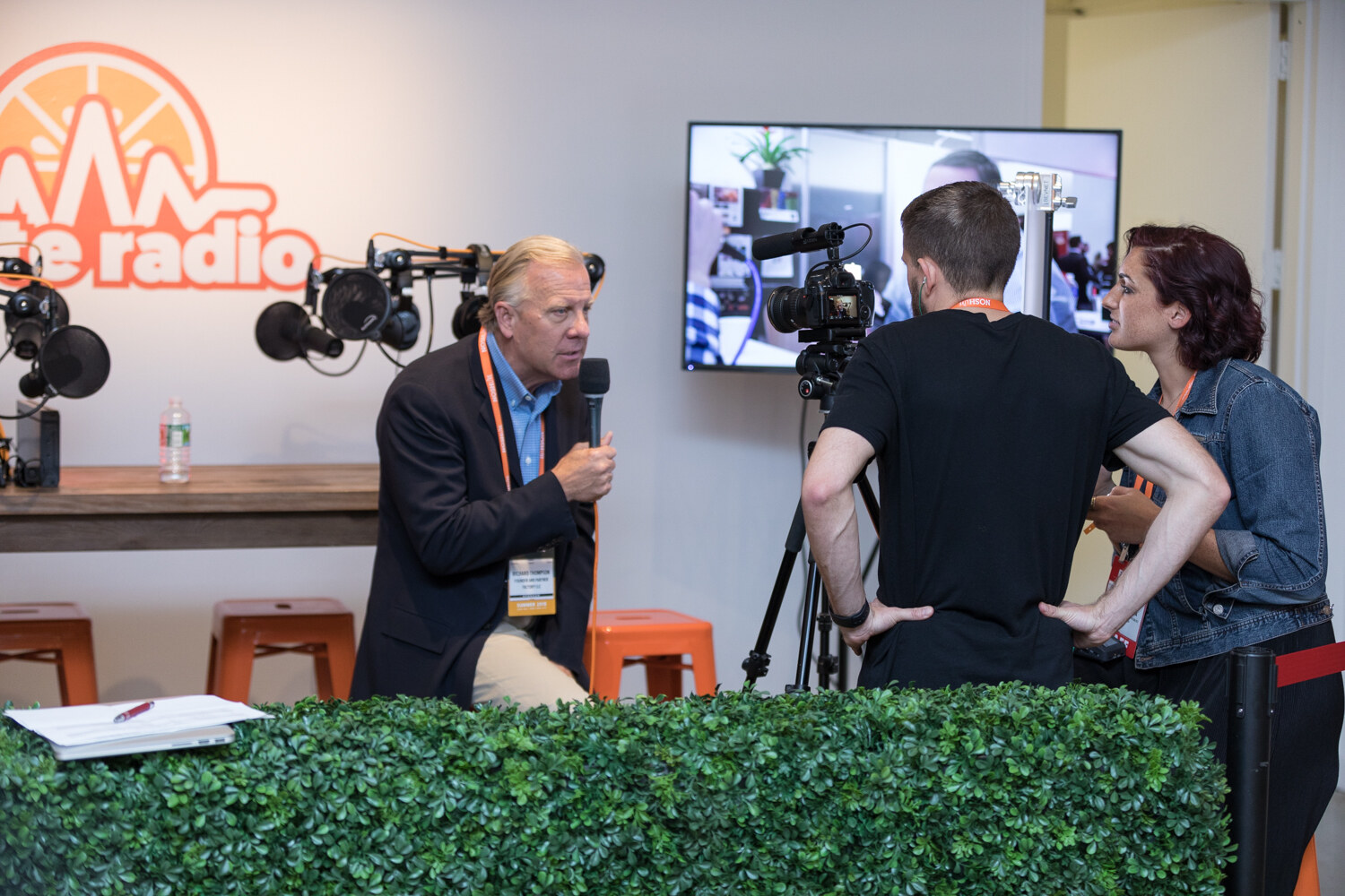 Two hosts and a camera operator record a Taste Radio livestream segment beside a Taste Radio sign and an orange stool.