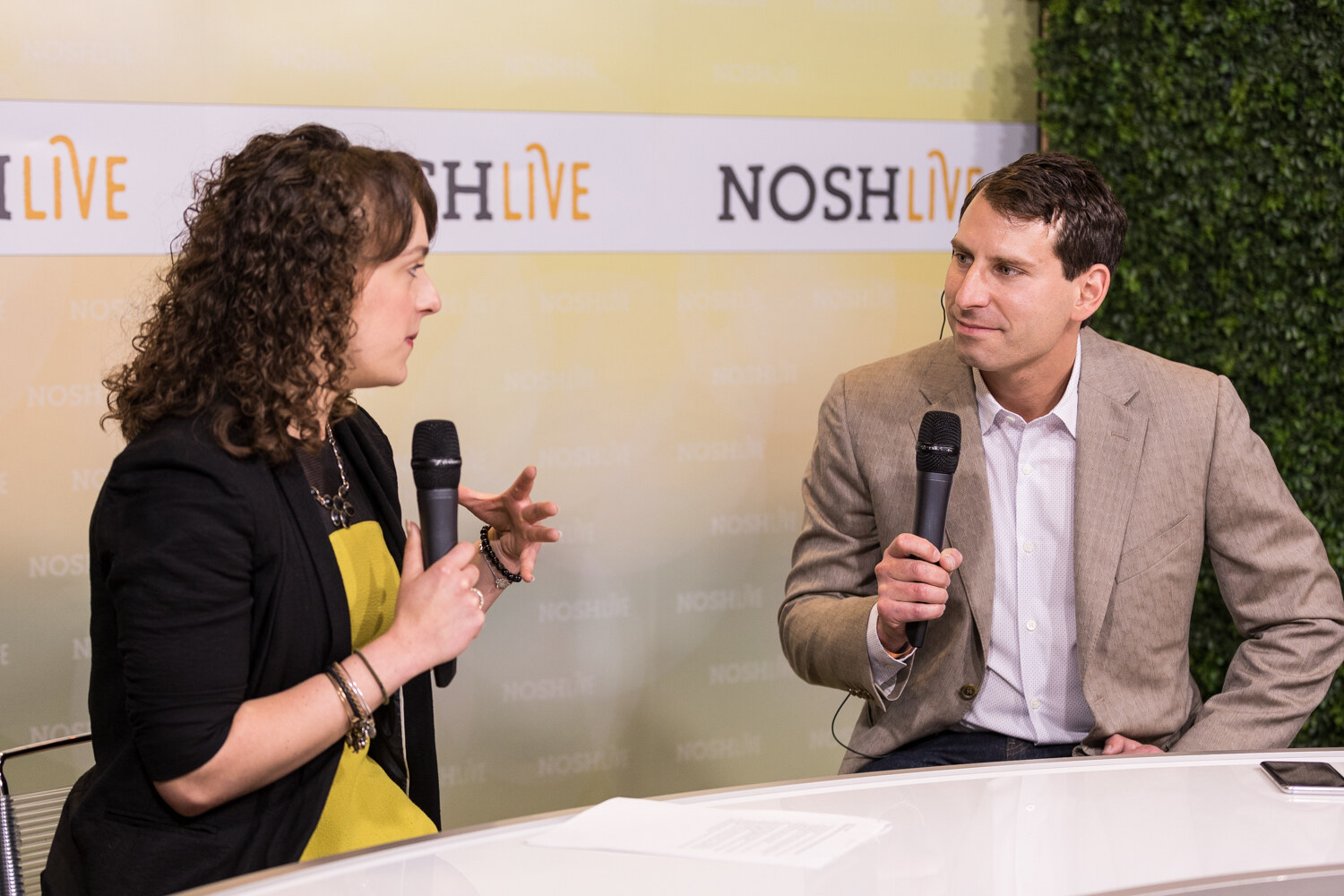 A host in a black blazer interviews a man in a tan blazer at the Nosh Live livestream desk.
