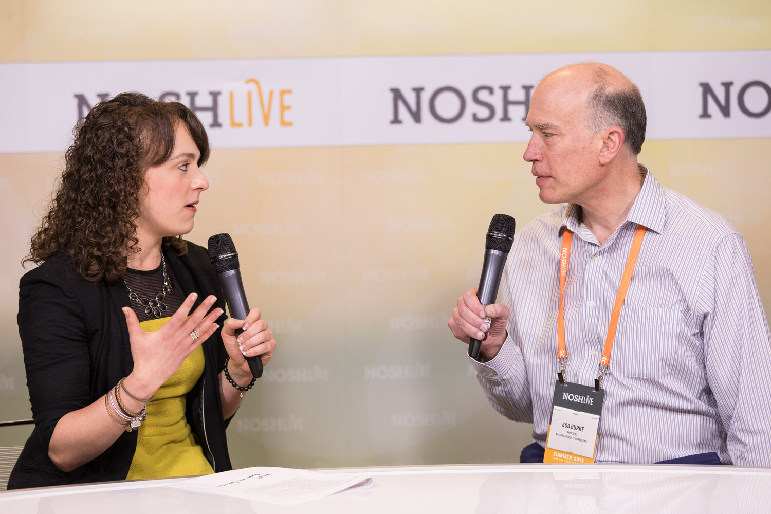 A host and a guest with a Nosh Live lanyard talk into microphones at the livestream desk.