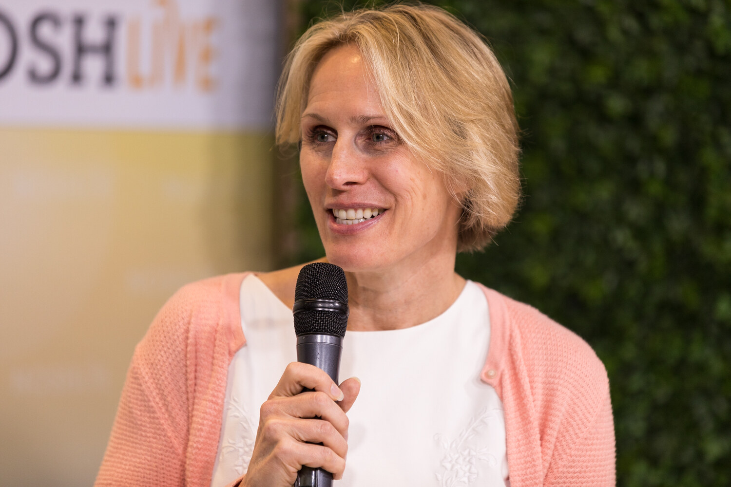 A livestream guest in a coral cardigan smiles while holding a microphone in front of an ivy wall.