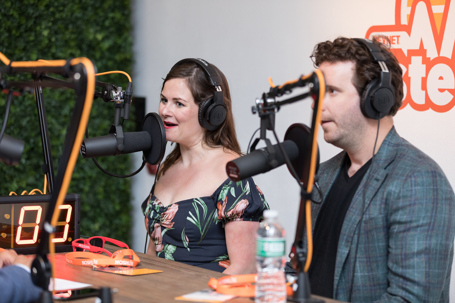 Two podcast hosts wearing headphones record at a table with microphone booms during Nosh Live.