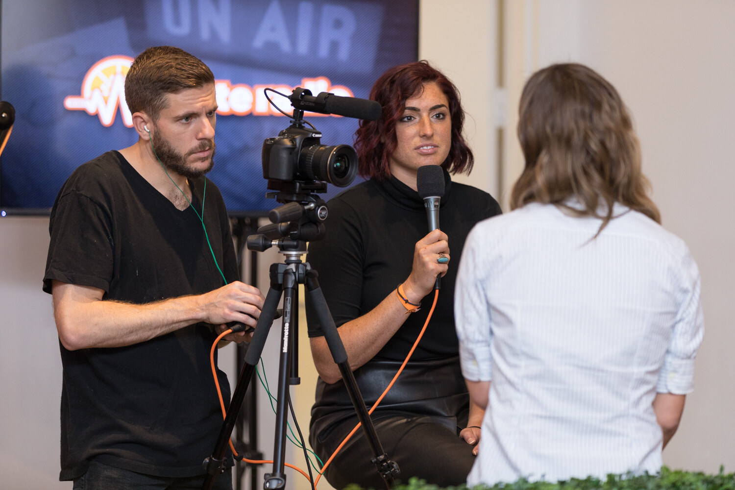 A camera operator and an interviewer hold a microphone beside a monitor reading On Air.