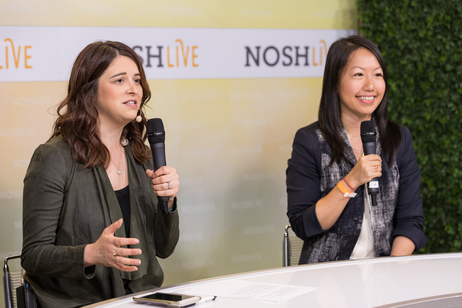 A host and a guest in a navy blazer talk into microphones at the Nosh Live livestream desk.
