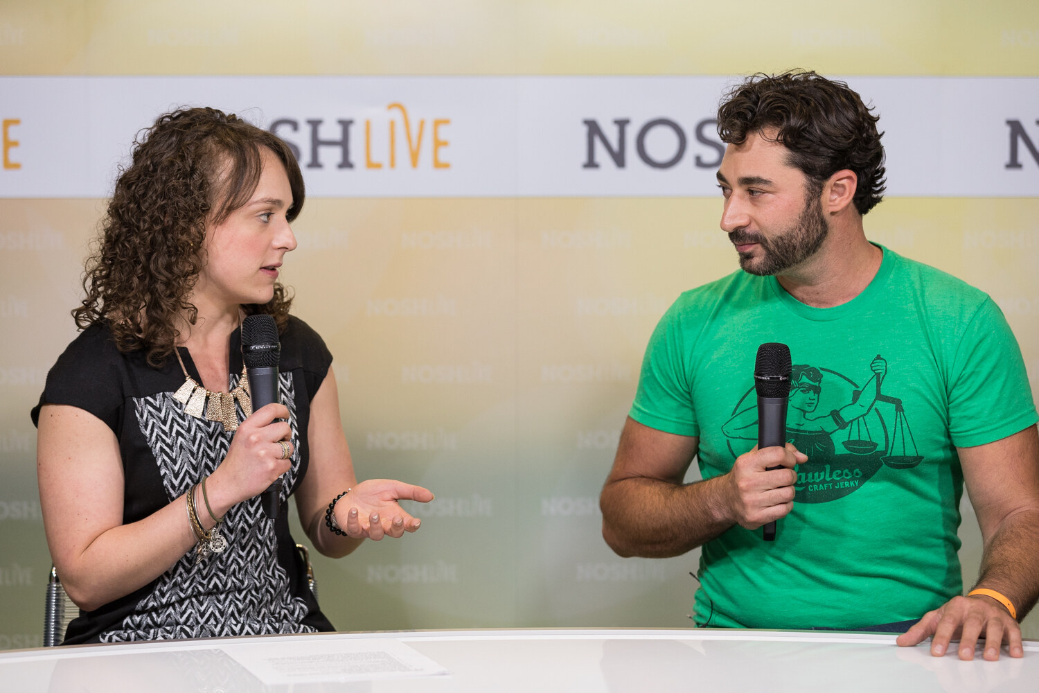 A host in a patterned top interviews a man in a green graphic t-shirt at the livestream desk.