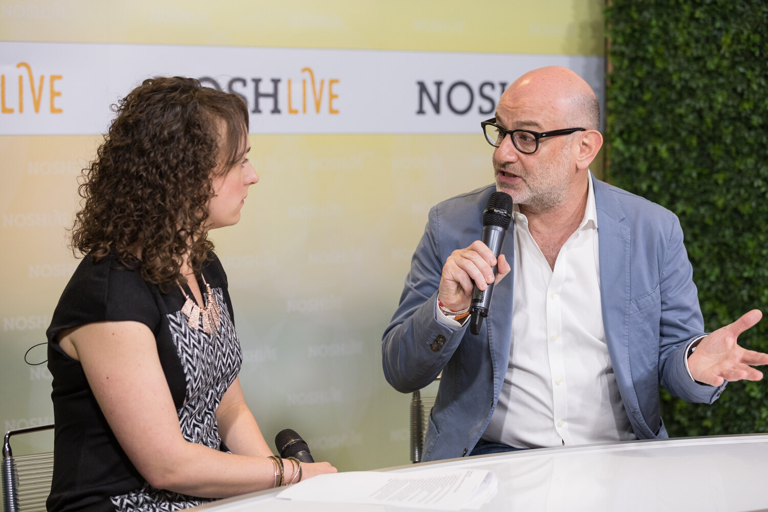 A livestream guest in a blue blazer and glasses gestures broadly while speaking to the host.
