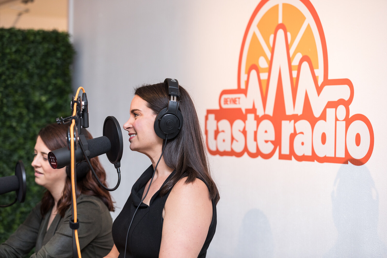 Two podcast hosts wearing headphones record at the Taste Radio corner with an orange and white Taste Radio logo wall.