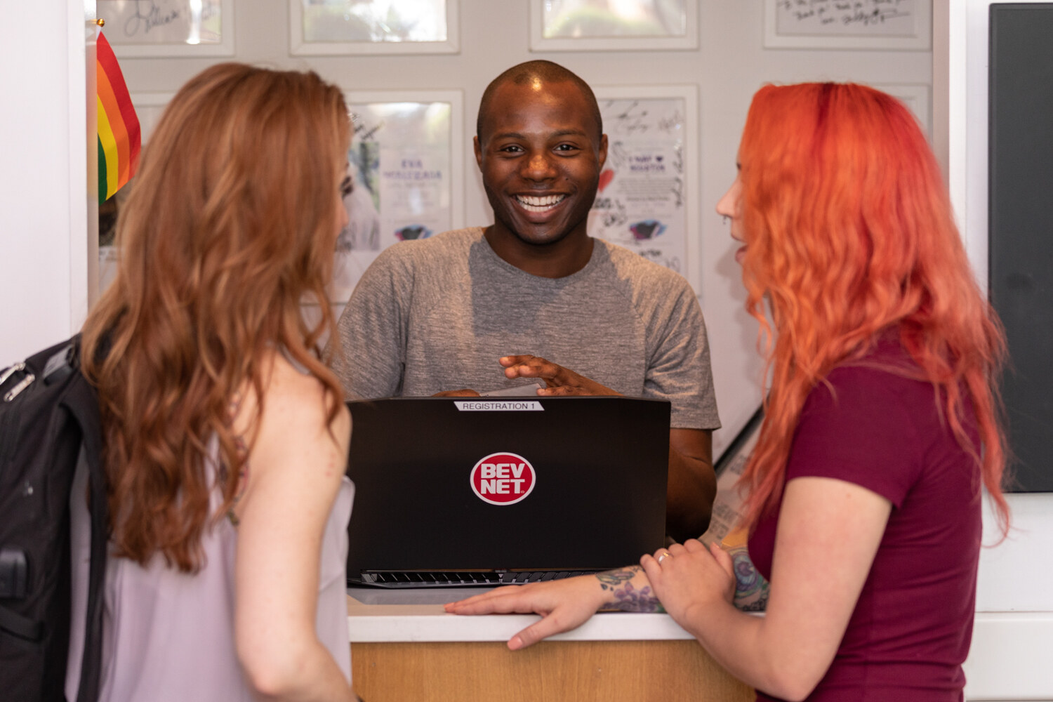 BEVNET registration staffer in a gray T-shirt smiling behind a laptop with a BEVNET sticker while greeting two women