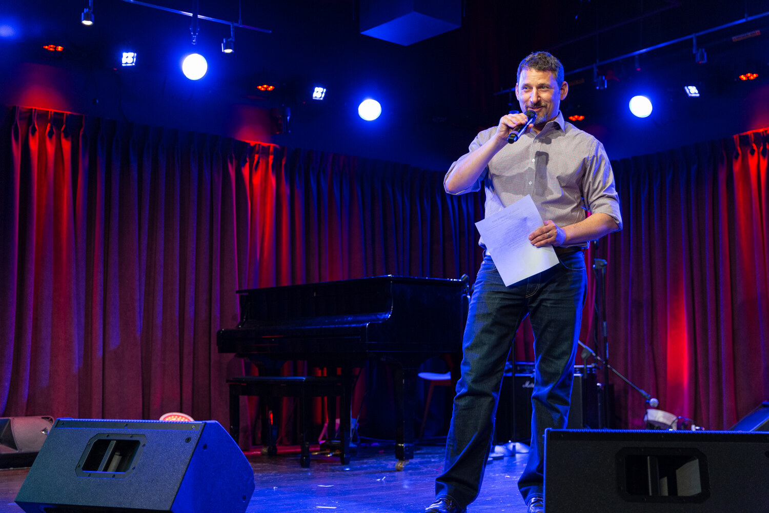 Boot Camp host in a gray checked shirt on stage holding a microphone and a sheet of paper