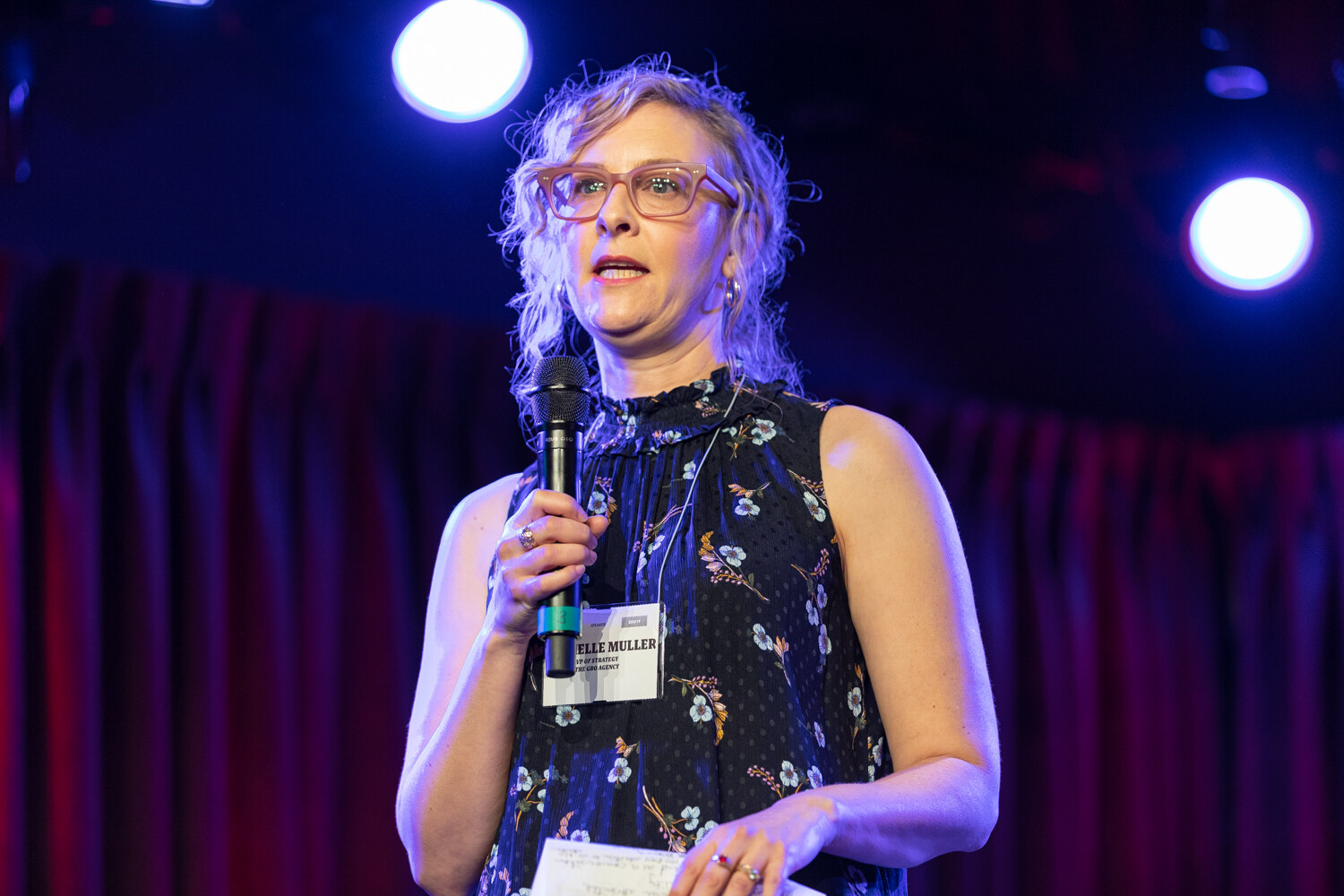 Boot Camp presenter with curly hair and red glasses in a navy floral sleeveless top holding a microphone