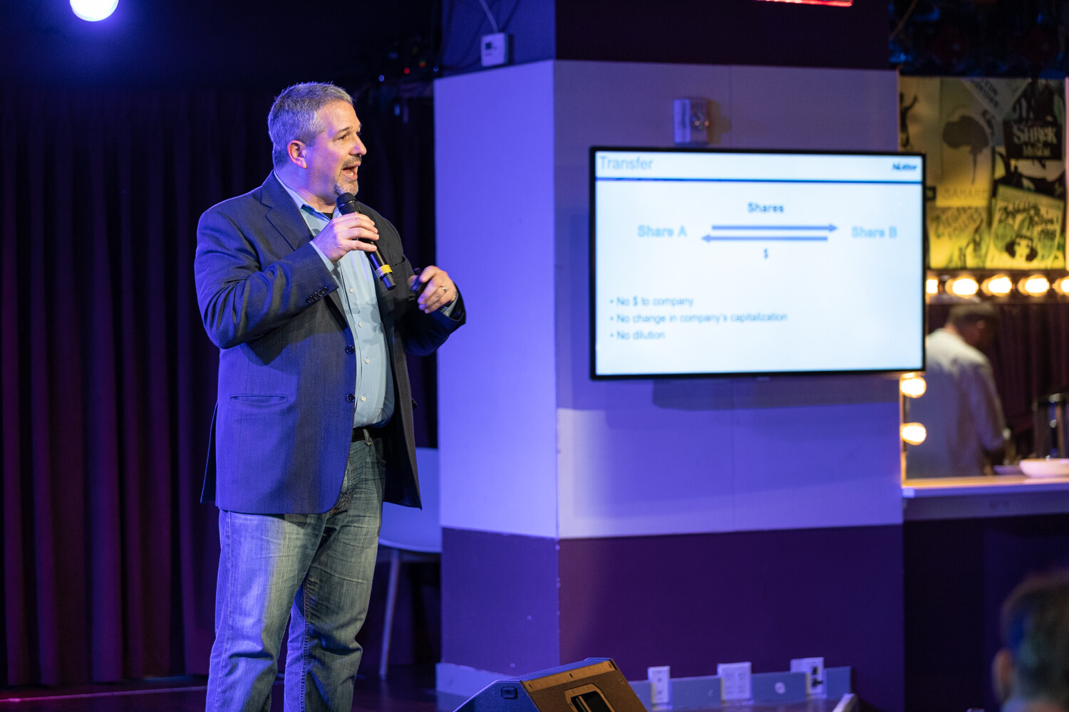 Boot Camp speaker in a navy blazer and jeans standing beside a wall screen showing a share transfer diagram