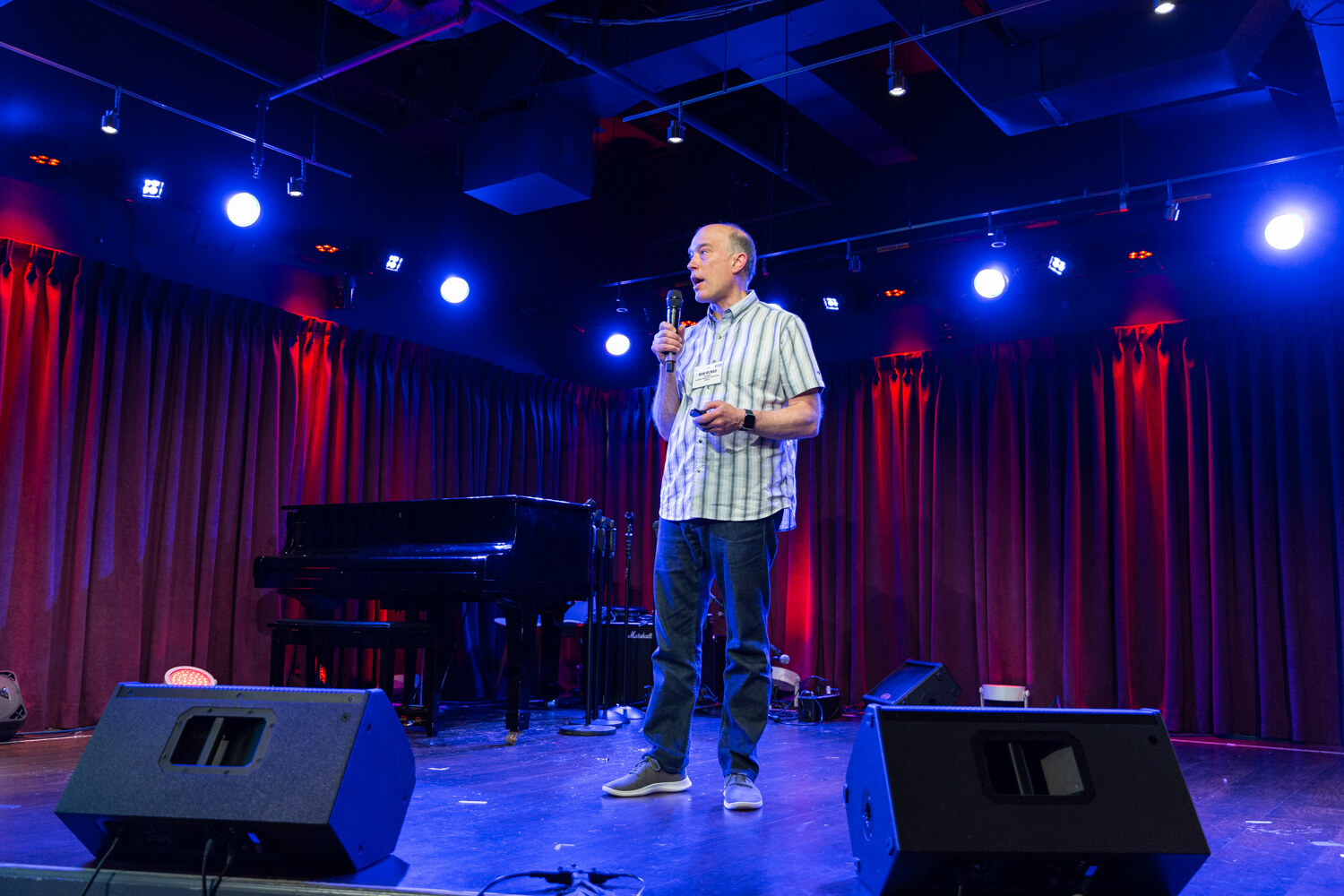 Wide view of the Boot Camp stage with the speaker standing beside a grand piano under blue lights