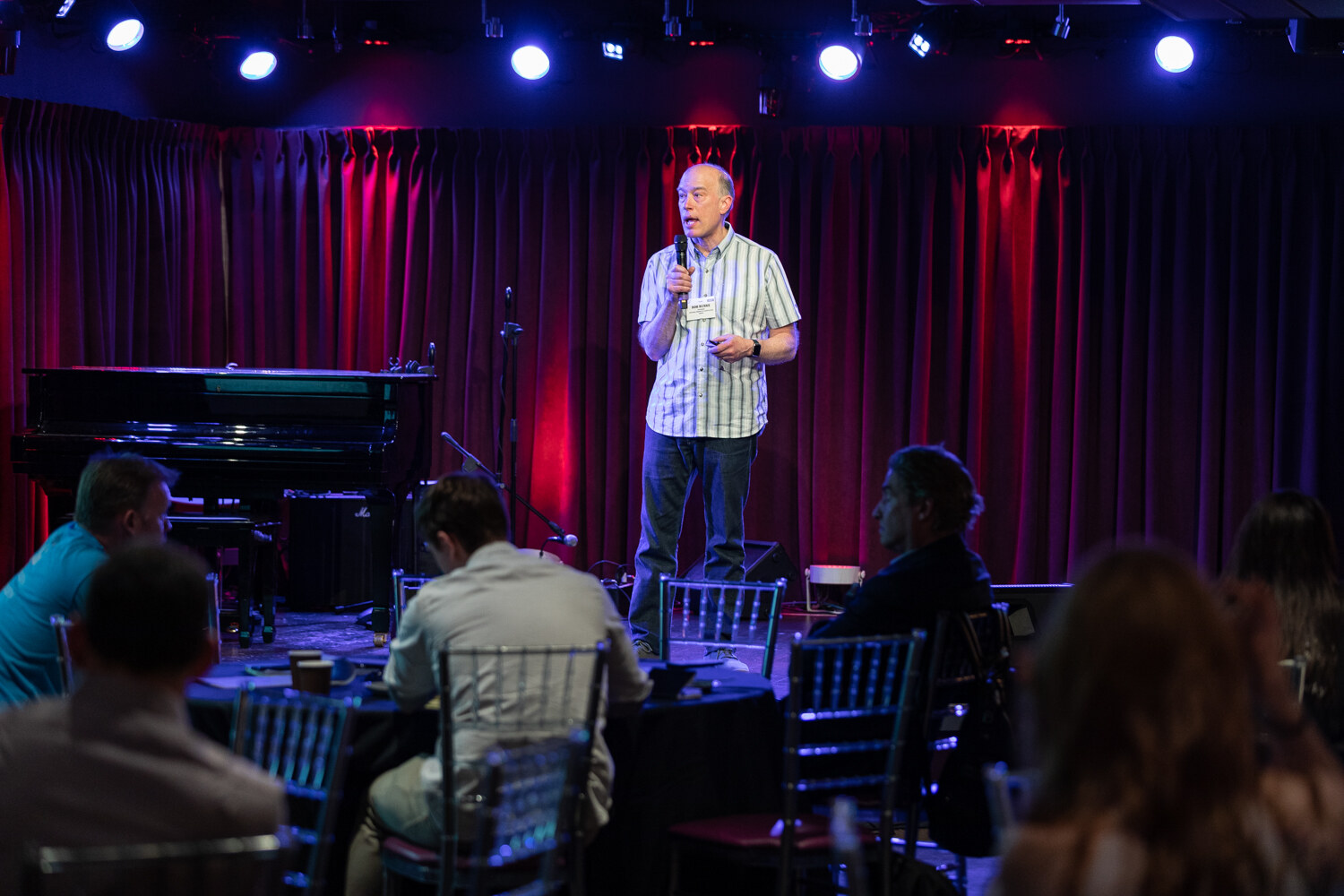 Boot Camp speaker standing on the red-curtained stage with attendees at tables in the foreground