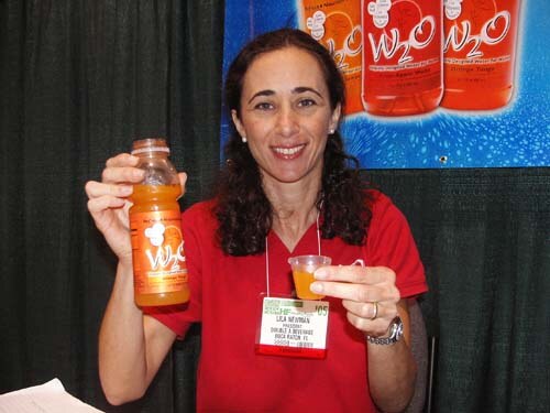 A smiling woman in a red shirt with a badge holds up a bottle of orange W2O in one hand and a small sample cup in the other, in front of W2O bottle graphics.