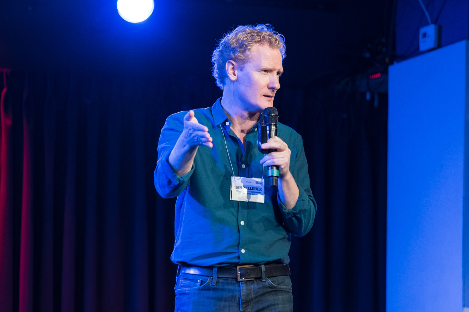 Boot Camp speaker with curly hair in a teal shirt gesturing toward the audience under blue stage light