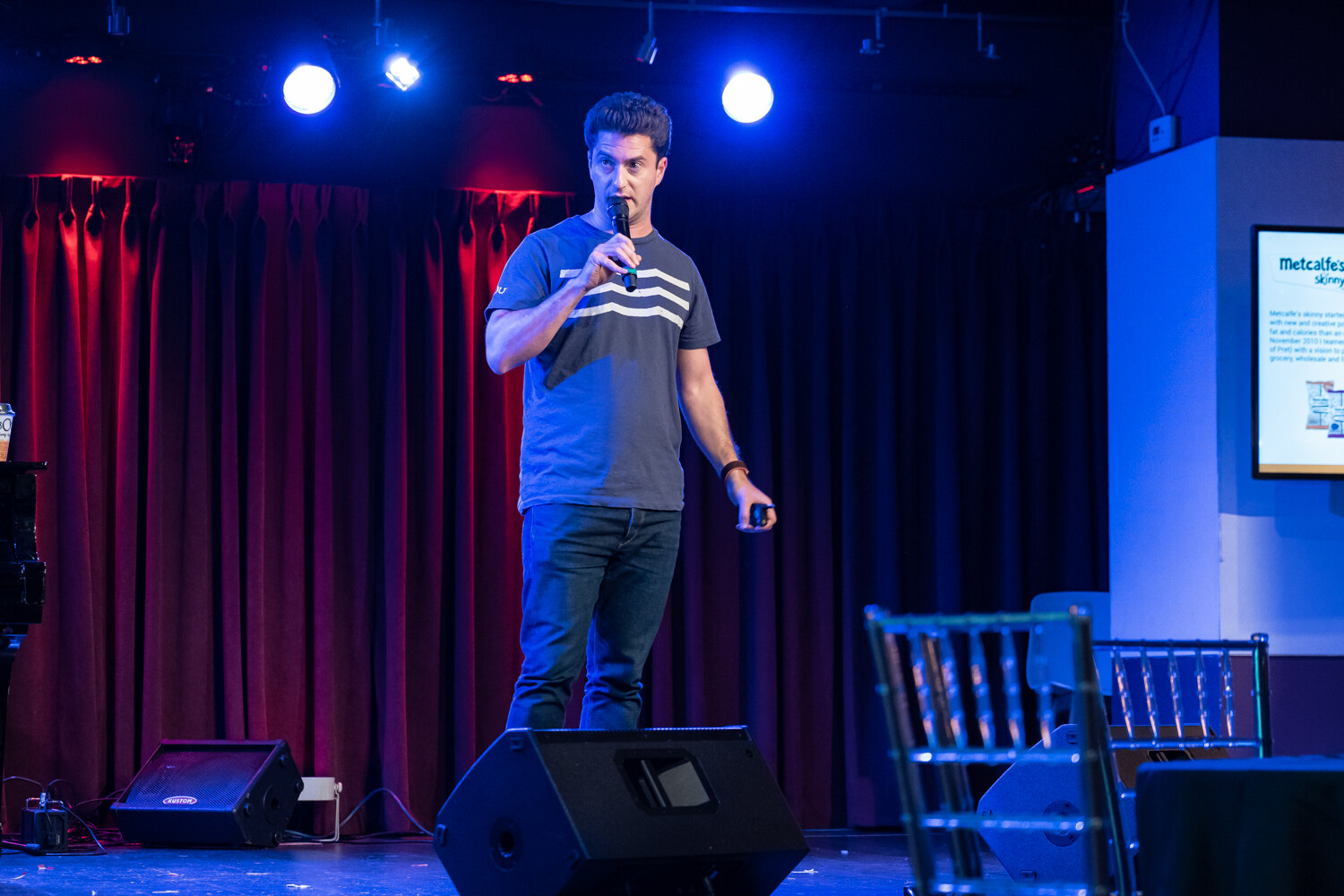 Boot Camp speaker in a gray T-shirt with white stripes standing on the blue-lit stage beside a Metcalfe's screen