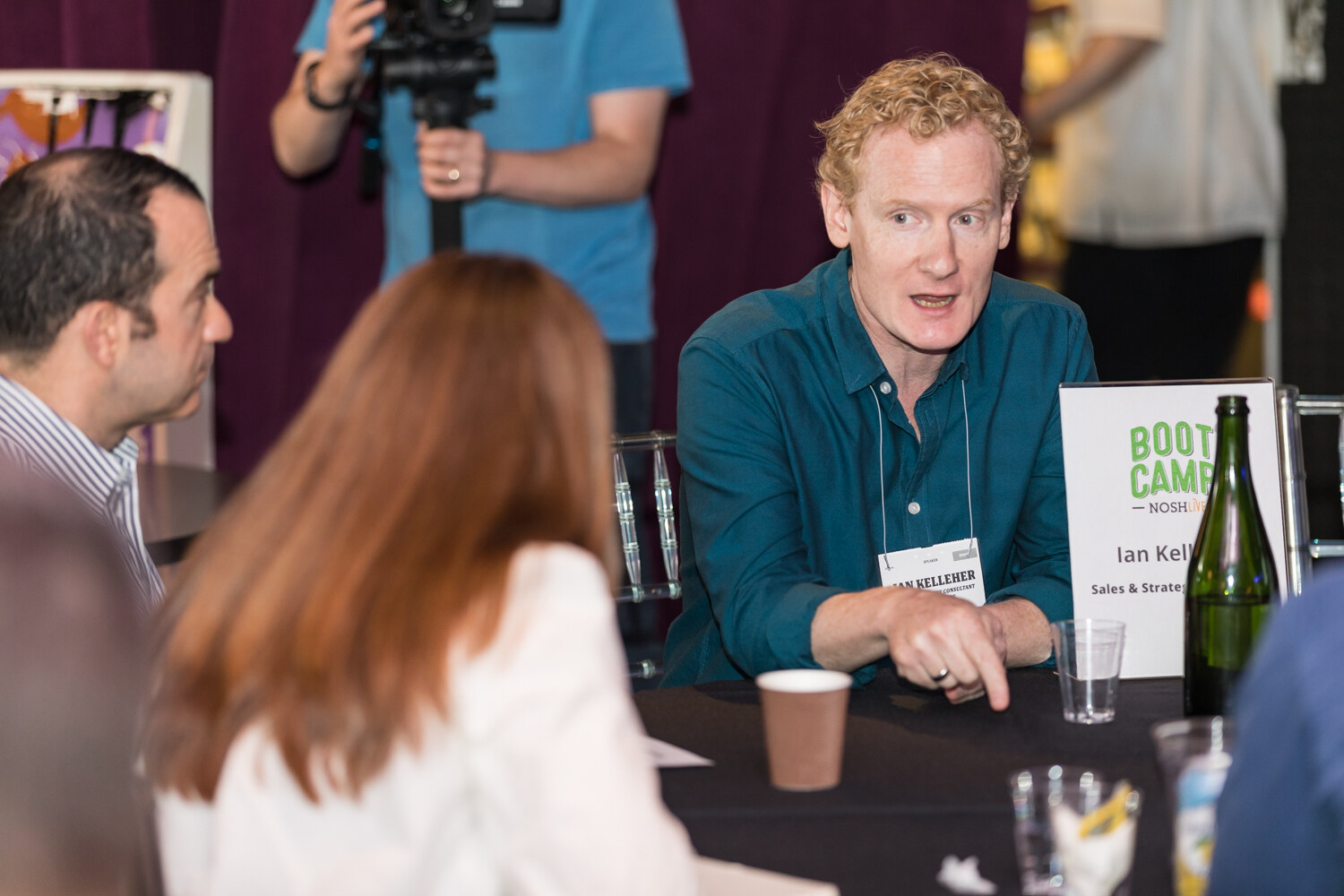Mentor with curly red hair in a teal shirt explaining something beside a Boot Camp table sign, a camera operator behind him