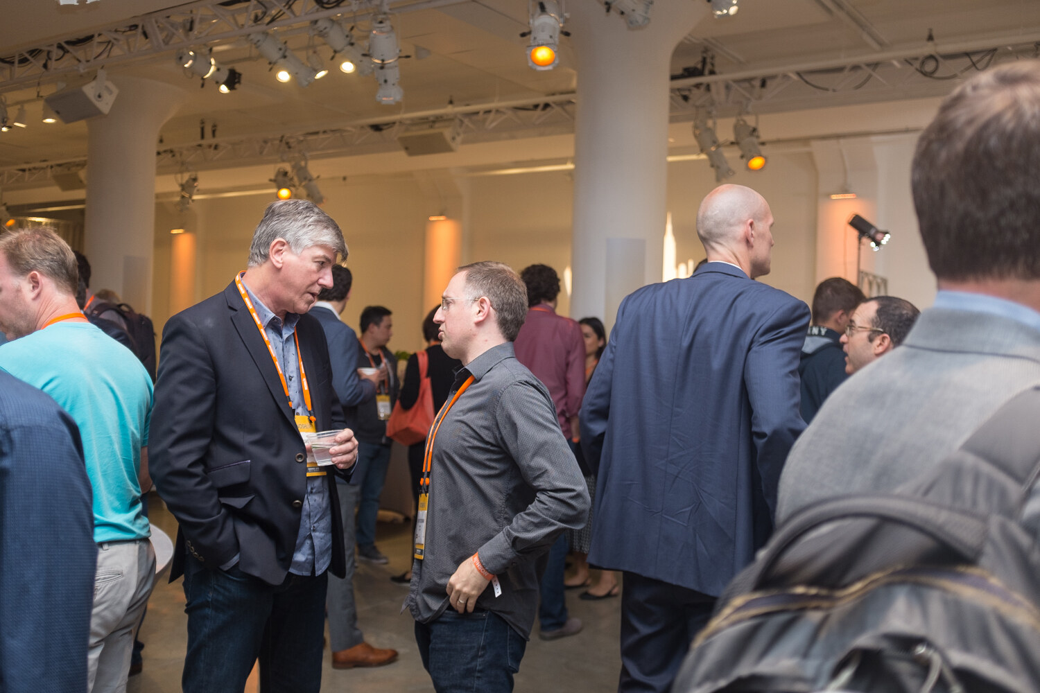 Crowded networking hall with attendees talking beneath white columns and warm lights