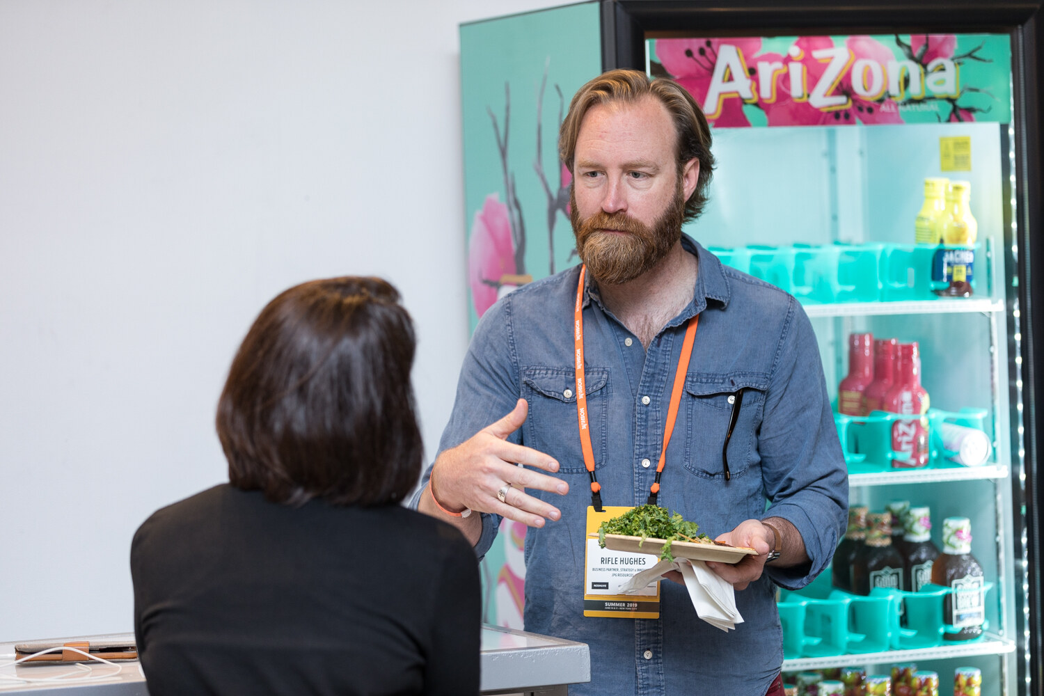 Bearded man in a denim shirt holding a plate and gesturing beside an AriZona refrigerator