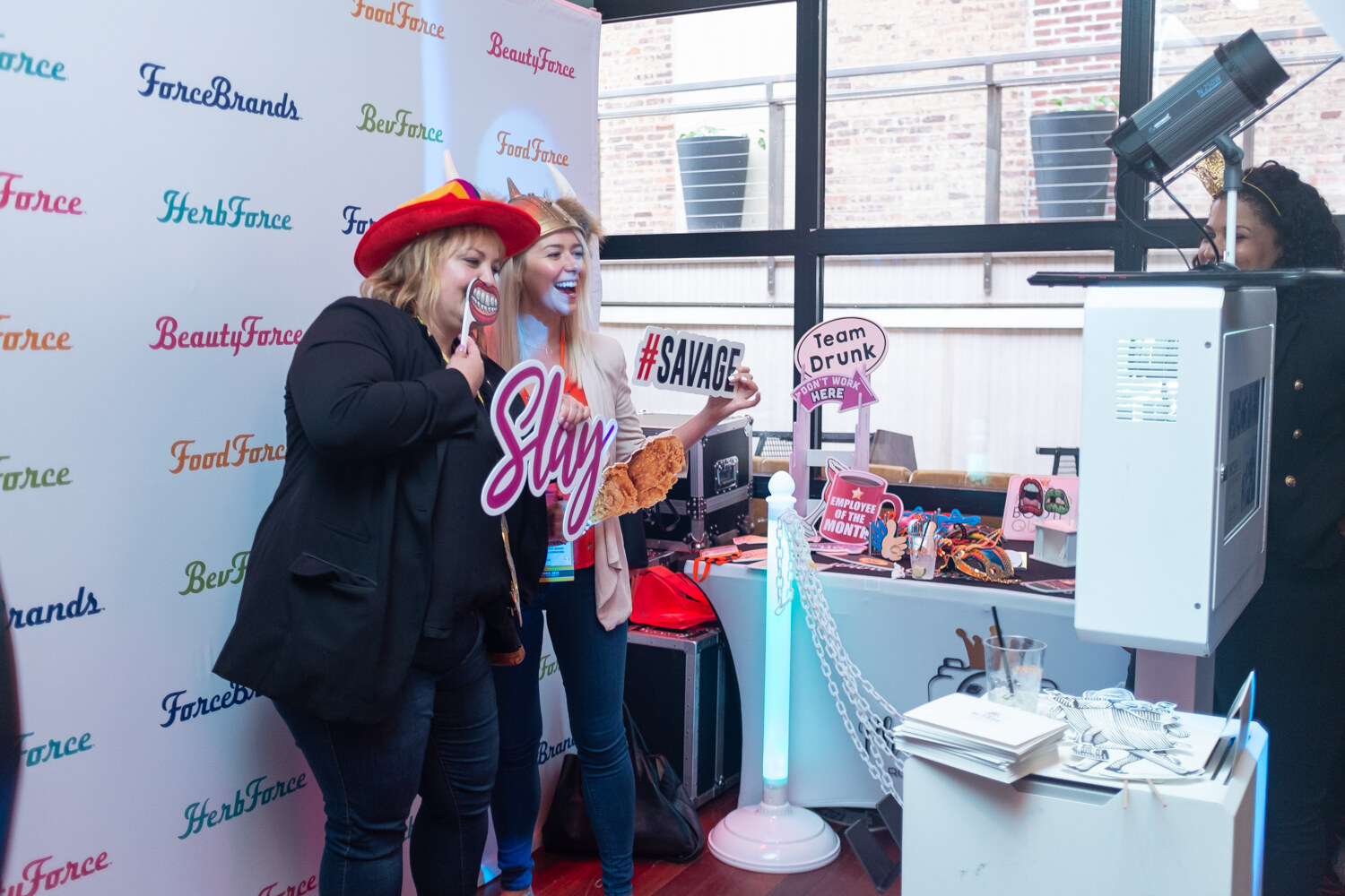 Two women wearing costume hats posing with props in front of the ForceBrands photo booth backdrop