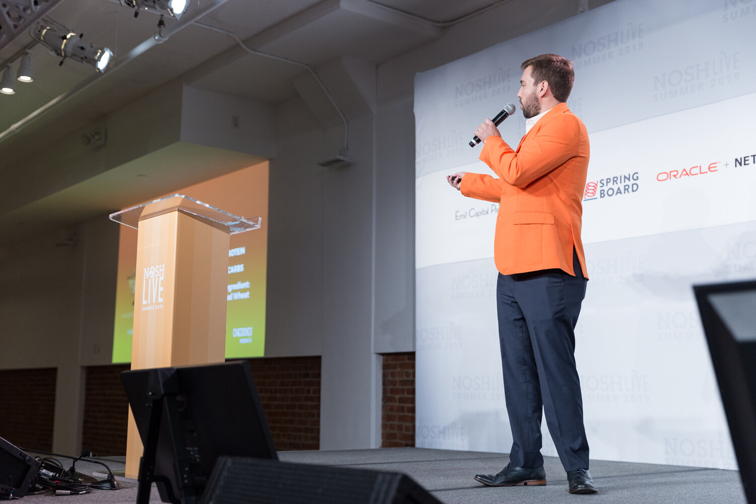 Pitch Slam presenter in an orange blazer speaking into a microphone on stage beside the lectern, seen from a low angle