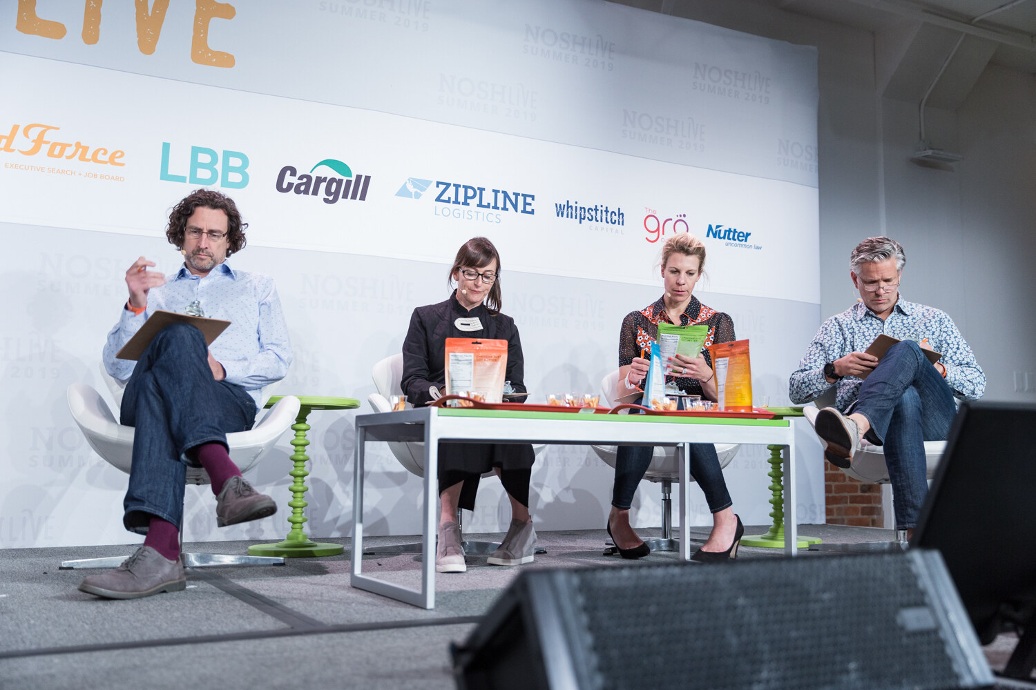 Four Pitch Slam judges seated on stage around a green table of snack pouches