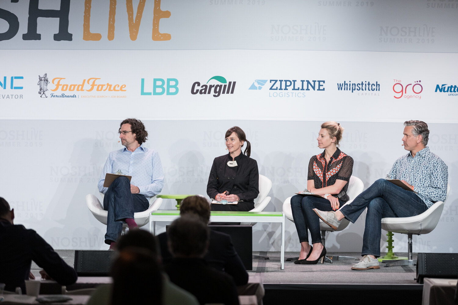 Four Pitch Slam judges seated on stage in front of the NOSHLIVE sponsor wall with audience heads in the foreground
