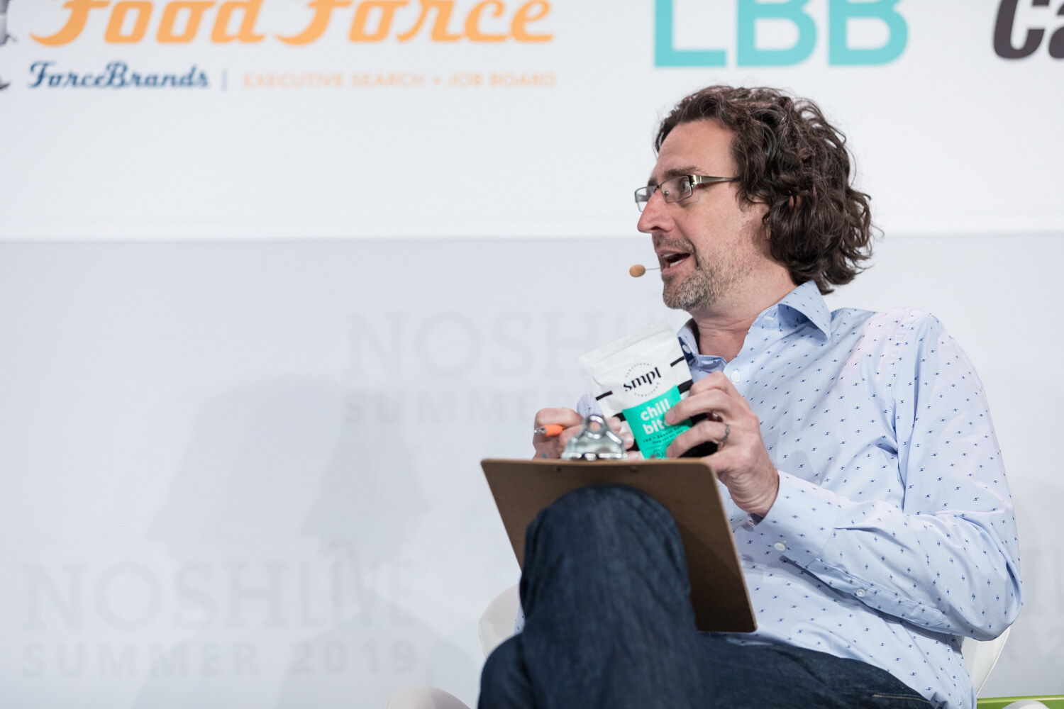 Curly-haired judge in a light blue shirt holding a Simpl chips bag while looking toward the stage