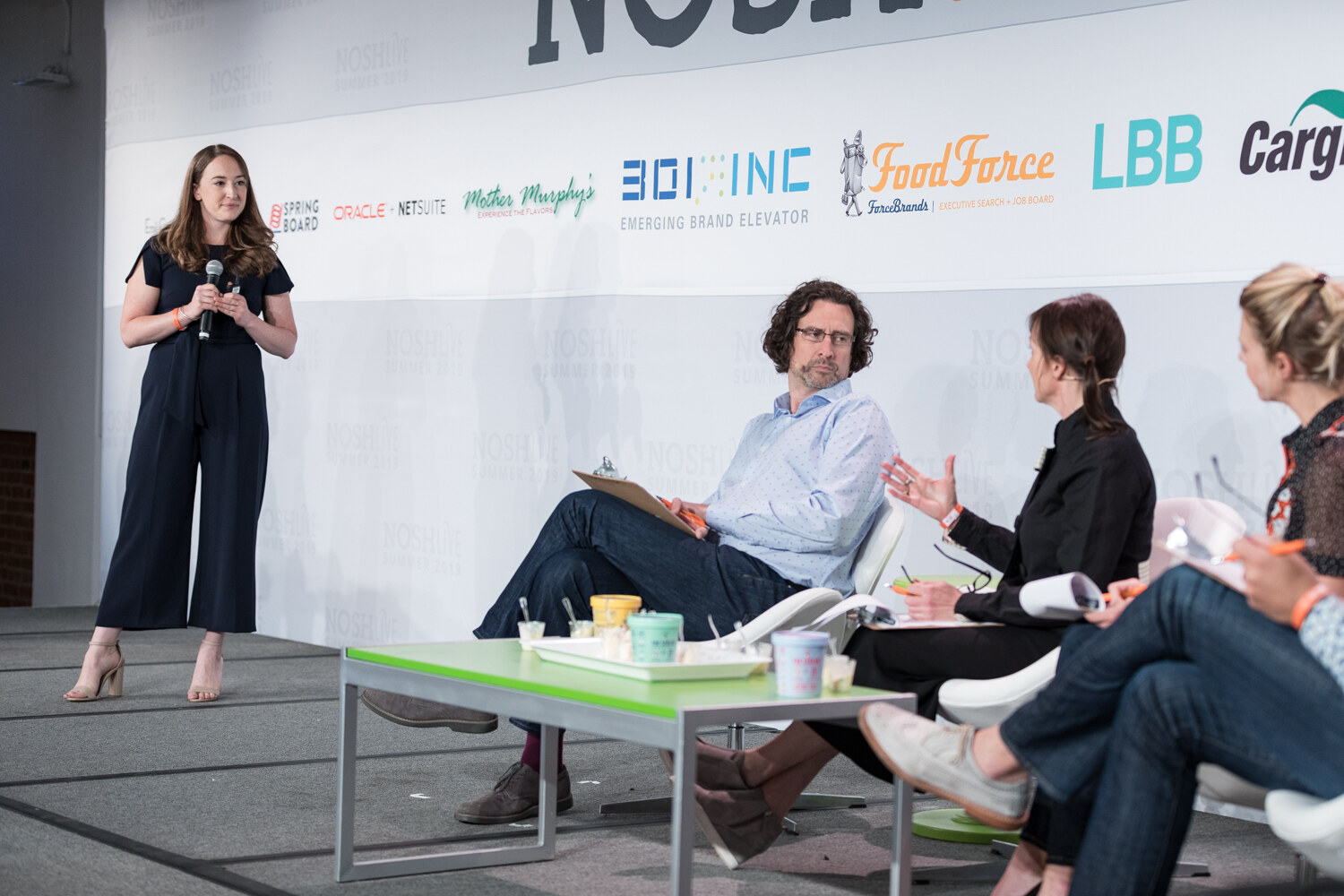Pitch Slam presenter in a navy jumpsuit standing beside four judges seated at a green table of ice cream tubs