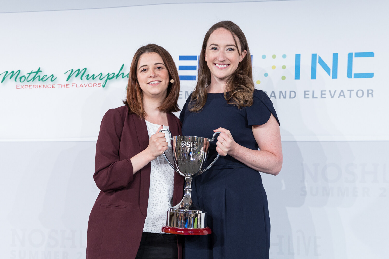 Two Pitch Slam winners, one in a maroon blazer and one in a navy jumpsuit, holding the trophy on stage