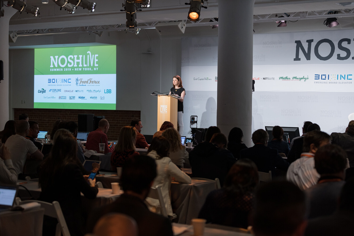 Wide view of the main hall with a green NOSH LIVE screen and a speaker at the lectern