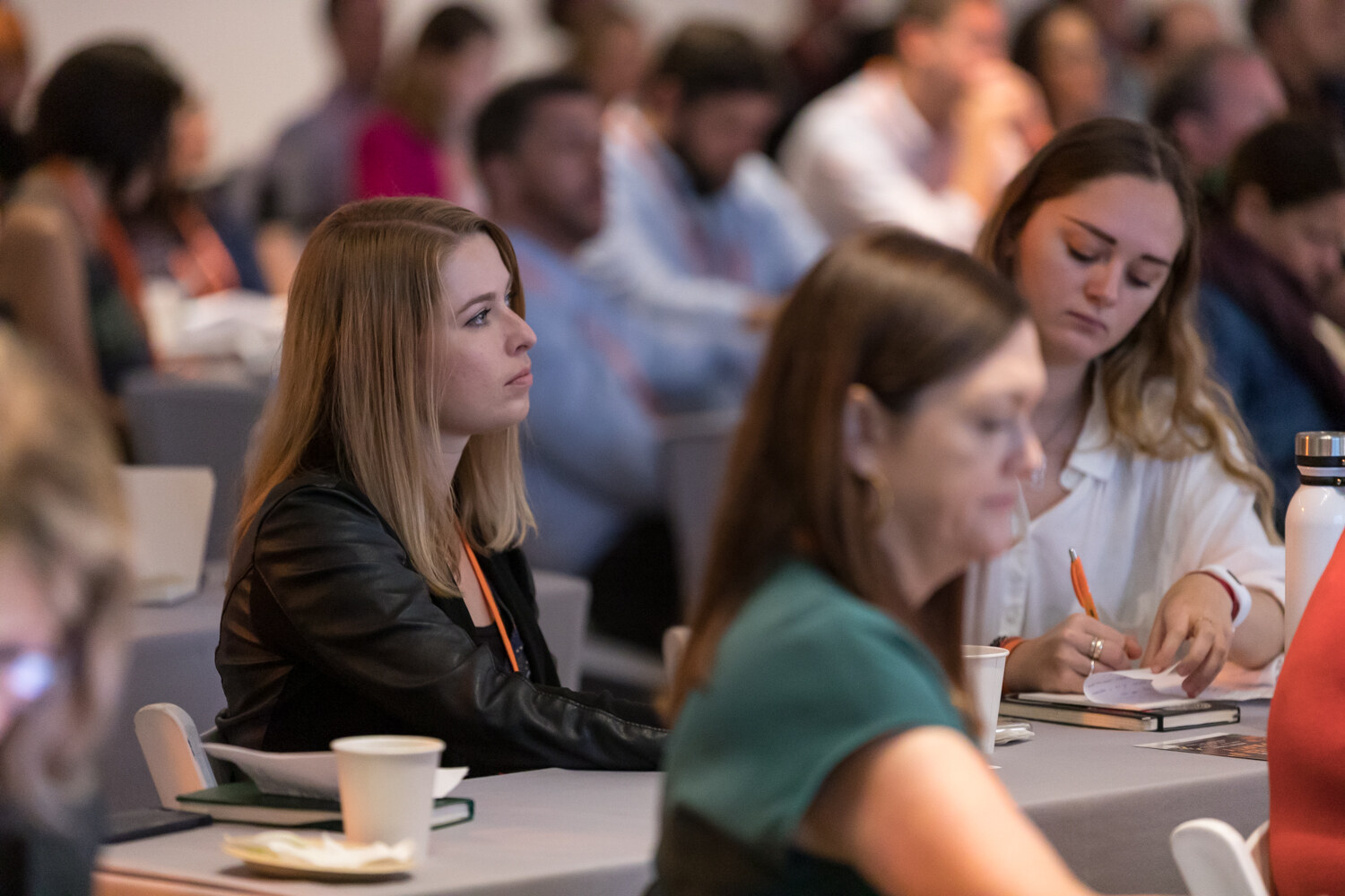 Attendees at draped tables listening to a session, a woman in a black leather jacket looking toward the stage