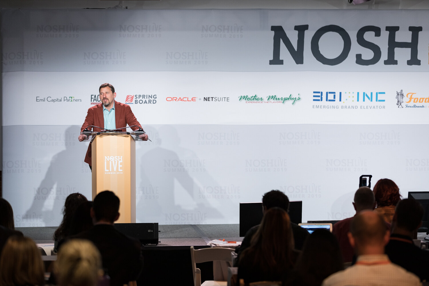 Host in a rust jacket at the NOSH LIVE lectern with audience heads in the foreground